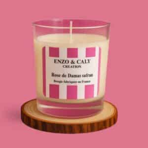 Bougie parfumée senteur Rose Damas safran, présentée dans un verre transparent avec étiquette rose et blanche, posée sur un rondin en bois et sur un fond uni rose – création élégante Enzo & Caly fabriquée en France.