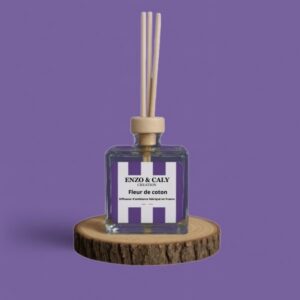 Diffuseur d’ambiance au parfum Fleur de coton, à tiges en bois, présenté dans un flacon carré en verre transparent, avec une étiquette mauve et blanche, posé sur un rondin en bois avec un fond uni mauve – création élégante Enzo & Caly fabriquée en France.