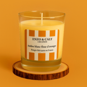 Bougie parfumée senteur Ambre blanc fleur d'oranger, présentée dans un verre transparent avec étiquette orange et blanche, posée sur un rondin en bois et sur un fond uni orange– création élégante Enzo & Caly fabriquée en France.