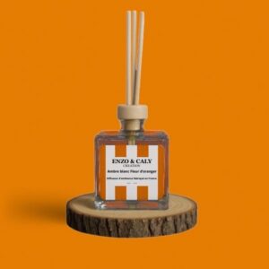 Diffuseur d’ambiance au parfum Ambre blanc fleur d'oranger, à tiges en bois, présenté dans un flacon carré en verre transparent, avec une étiquette orange et blanche, posé sur un rondin en bois avec un fond uni orange – création élégante Enzo & Caly fabriquée en France.