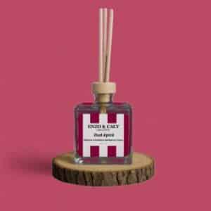 Diffuseur d’ambiance au parfum Oud épicé, à tiges en bois, présenté dans un flacon carré en verre transparent, avec une étiquette bordeaux et blanche, posé sur un rondin en bois avec un fond uni bordeaux – création élégante Enzo & Caly fabriquée en France.