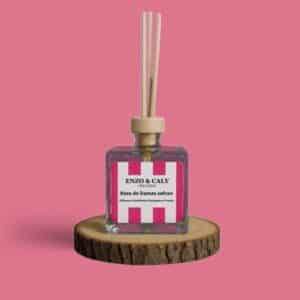 Diffuseur d’ambiance au parfum Rose Damas safran, à tiges en bois, présenté dans un flacon carré en verre transparent, avec une étiquette rose et blanche, posé sur un rondin en bois avec un fond uni rose – création élégante Enzo & Caly fabriquée en France.