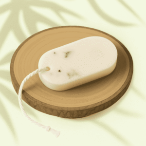 Tablette de cire parfumée au sakura René et Gigi, de forme ovale, de couleur crème, à ficelle, incrustée de petits fragments végétaux verts, exposée sur une base en bois dans une ambiance lumineuse naturelle.