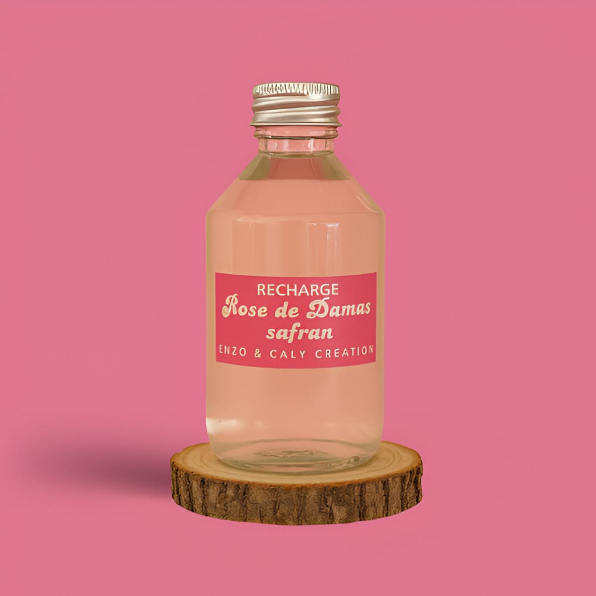 Recharge de parfum d’ambiance Rose Damas safran, présentée dans une bouteille en verre transparent avec étiquette rose, posée sur un rondin en bois et un fond uni rose – création élégante Enzo & Caly fabriquée en France.
