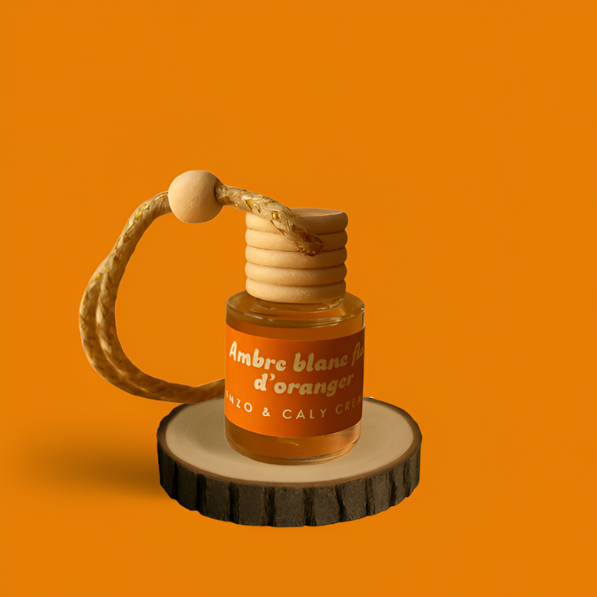 Mini diffuseur au parfum Ambre blanc fleur d'oranger, présenté dans un petit flacon en verre transparent, avec une étiquette orange et un cordon torsadé, posé sur un rondin en bois avec un fond uni orange – création élégante Enzo & Caly fabriquée en France.