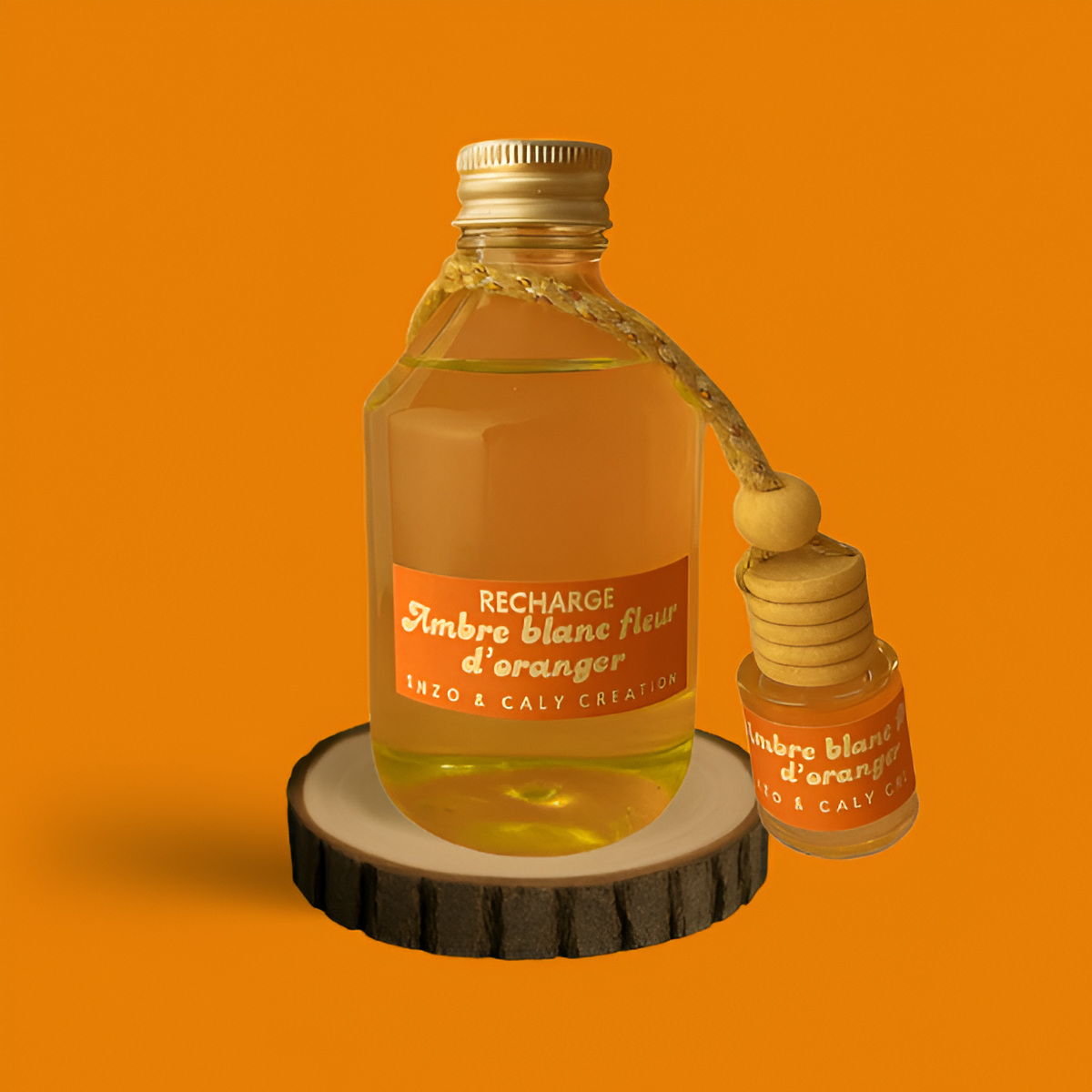 Ensemble composé d’un mini diffuseur de parfum d'ambiance Ambre blanc fleur d'oranger et de sa recharge, présentés respectivement dans un petit flacon et dans une bouteille en verre transparent, avec une étiquette orange, posés sur un rondin en bois avec un fond uni orange – création élégante Enzo & Caly fabriquée en France.