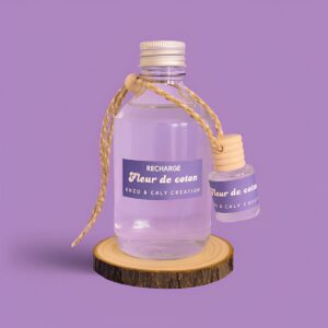 Ensemble composé d’un mini diffuseur de parfum d'ambiance Fleur de coton et de sa recharge, présentés respectivement dans un petit flacon et dans une bouteille en verre transparent, avec une étiquette mauve, posés sur un rondin en bois avec un fond uni mauve – création élégante Enzo & Caly fabriquée en France.