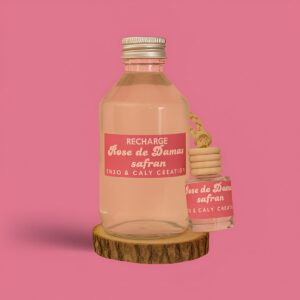 Mini diffuseur au parfum Rose Damas safran, présenté dans un petit flacon en verre transparent, avec une étiquette rose et un cordon torsadé, posé sur un rondin en bois avec un fond uni rose – création élégante Enzo & Caly fabriquée en France.