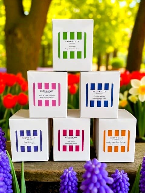 Six Bougies parfumées en boîtes cartonnées colorées, rose, bleu, bleu-mauve, pourpre et orange, disposées en pyramide et présentées en extérieur dans un jardin fleuri avec tulipes, narcisses et jacinthes. »– créations élégantes Enzo & Caly fabriquée en France.